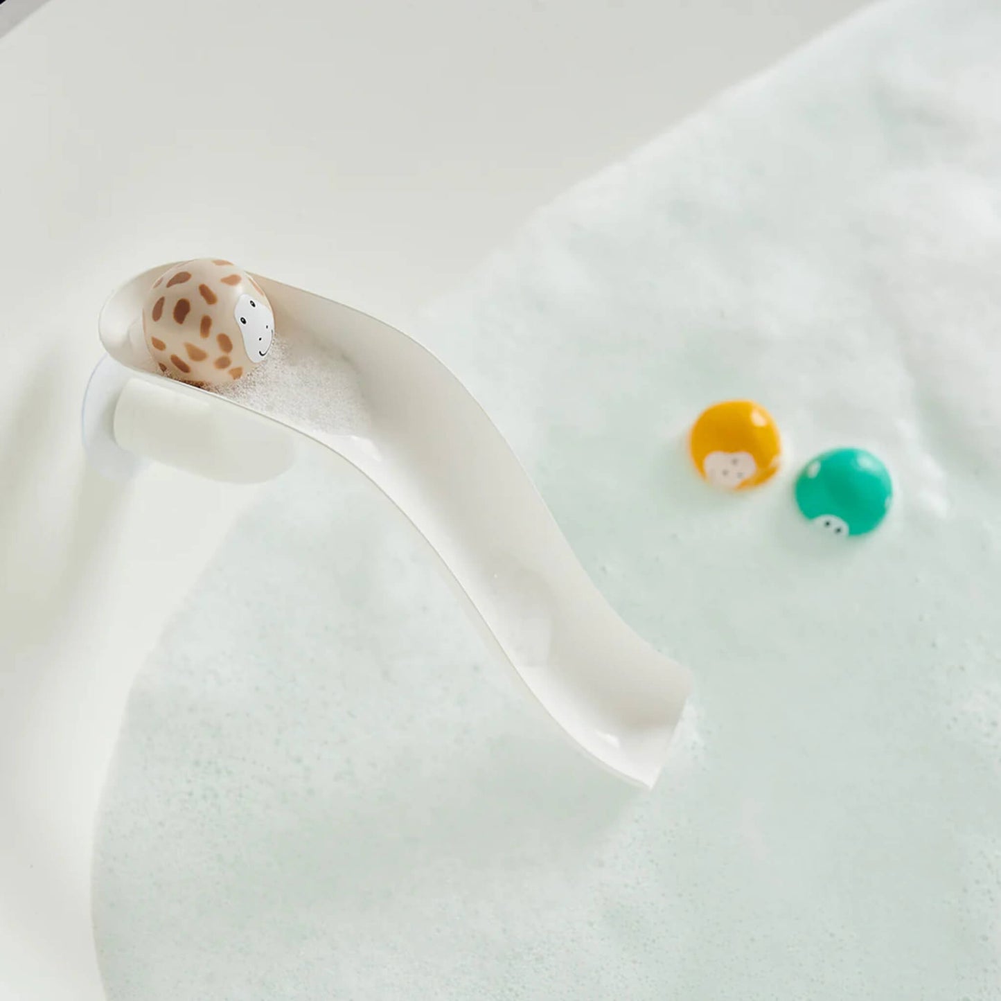 White curved slide attached to the bath side with a beige animal ball resting at the top, sitting above a surface of foamy bath water.
