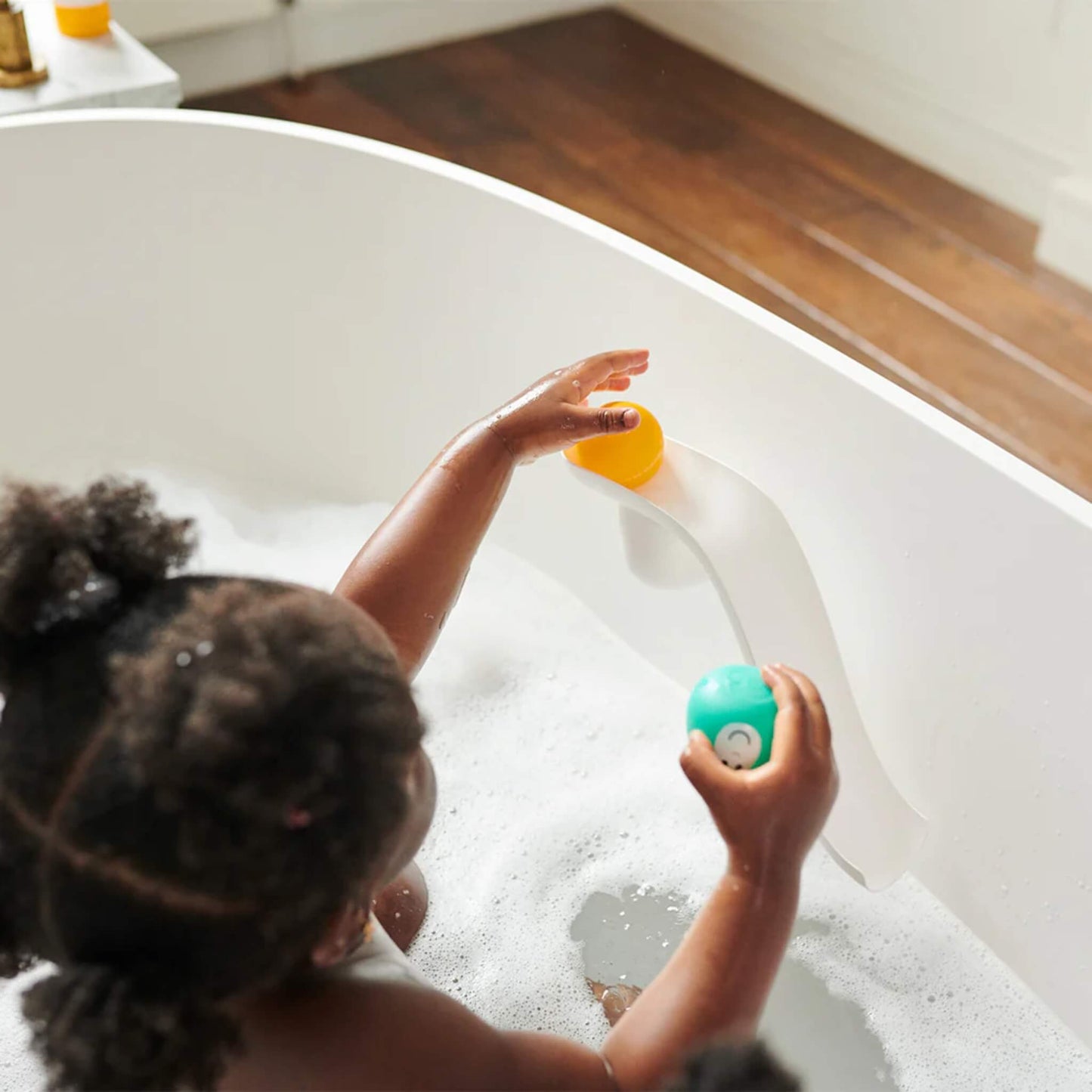 Child sitting in a bubble-filled bath while placing a yellow animal ball onto a white curved slide attached to the bath wall.