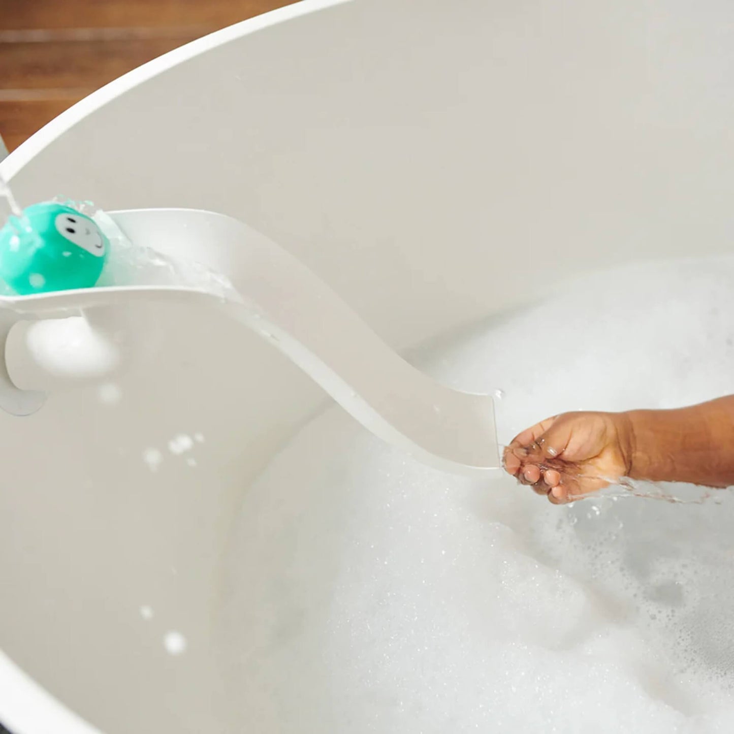 White curved slide attached to the bath wall with a turquoise animal ball rolling down as water flows from the top, with a child’s hand catching the stream.