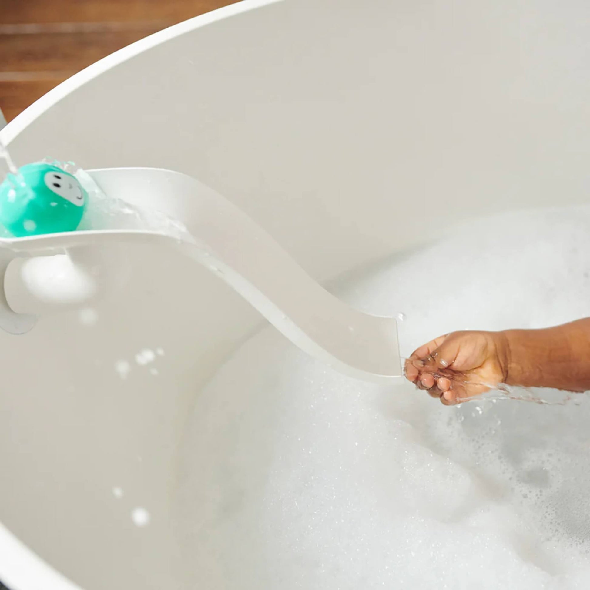 White curved slide attached to the bath wall with a turquoise animal ball rolling down as water flows from the top, with a child’s hand catching the stream.