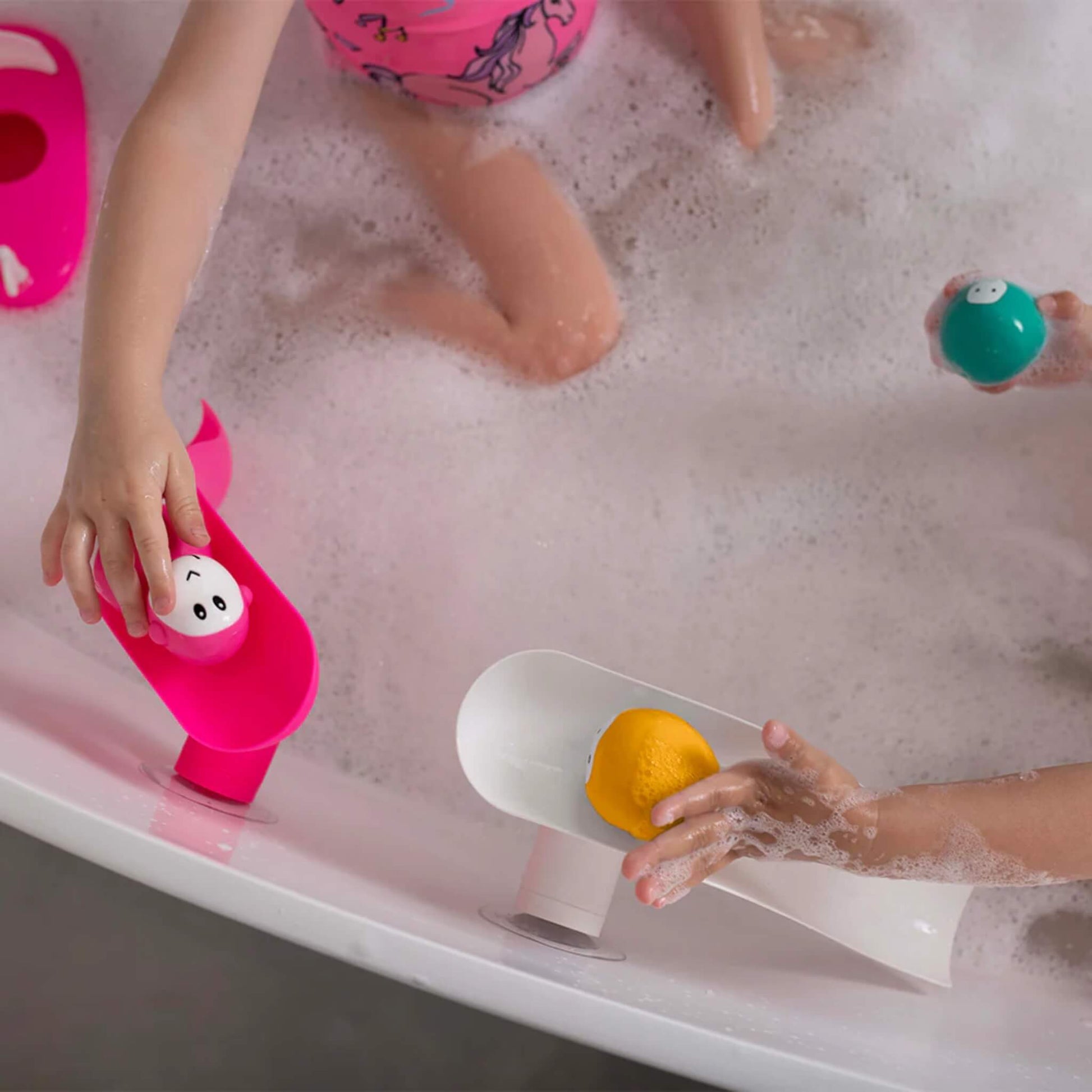 Two children playing in a foamy bath with a white slide and a pink slide attached to the bath rim, each rolling animal balls down their own slide.