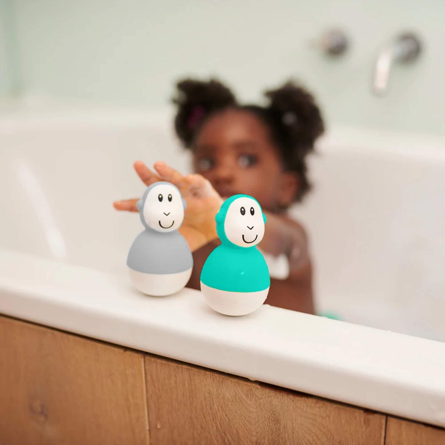 A young child reaching toward two wobbling bath toys resting on the edge of a bathtub, with soft bathroom lighting in the background.