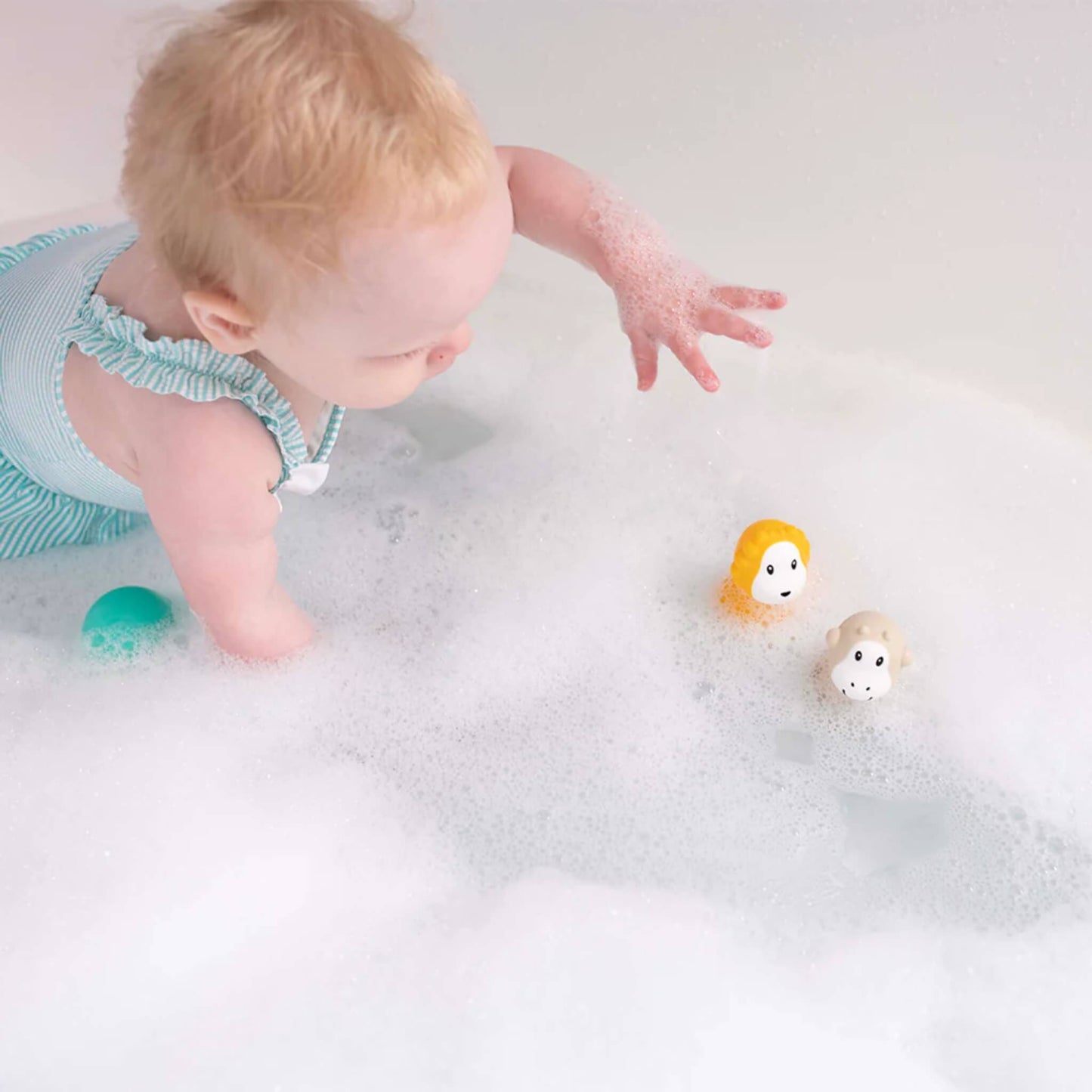 A baby reaching through bath bubbles toward two wobbling bath toys floating on the water.