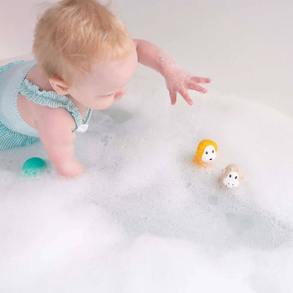 A baby reaching through bath bubbles toward two wobbling bath toys floating on the water.
