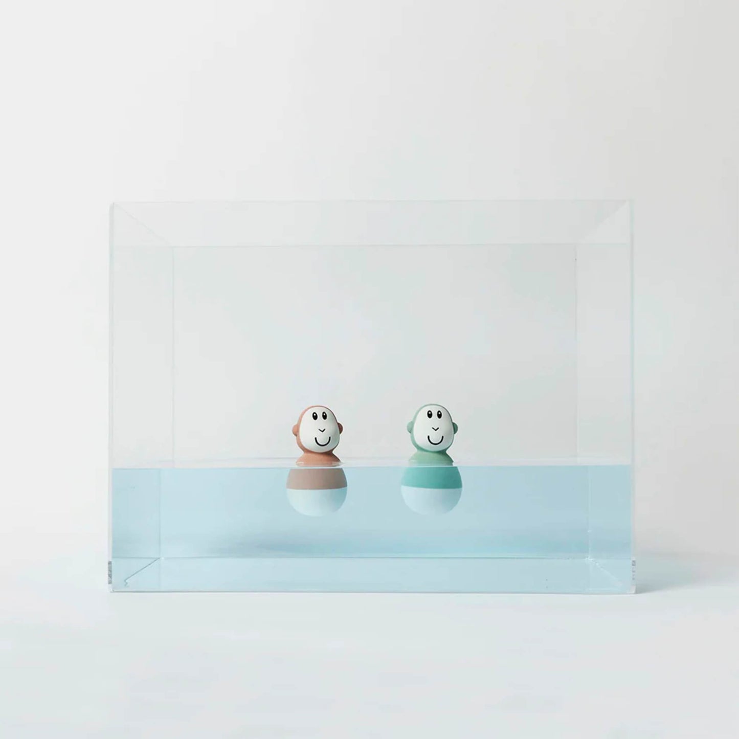 Two pastel-coloured wobbling bath toys float upright in a clear acrylic tank filled halfway with pale blue water.