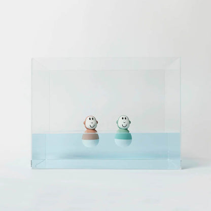 Two pastel-coloured wobbling bath toys float upright in a clear acrylic tank filled halfway with pale blue water.