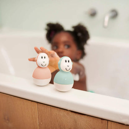 A toddler reaches toward two pastel-coloured wobbling bath toys placed on the edge of a tub, with soft lighting and bathroom tiles in the background.