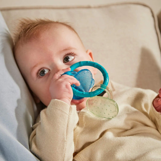 A baby lying on a soft beige cushion holding and chewing a turquoise silicone teething ring with attached blue and clear monkey face cooling teethers.