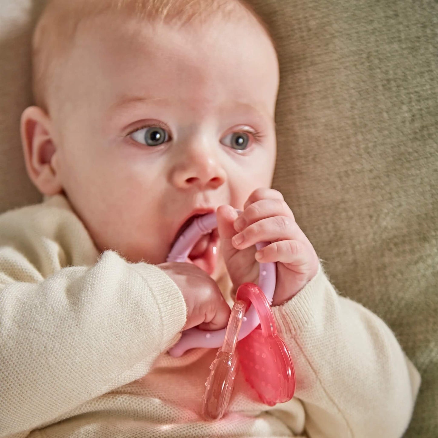A baby sitting against a soft cushion chewing a lilac silicone teething ring with attached pink and clear monkey face cooling teethers.