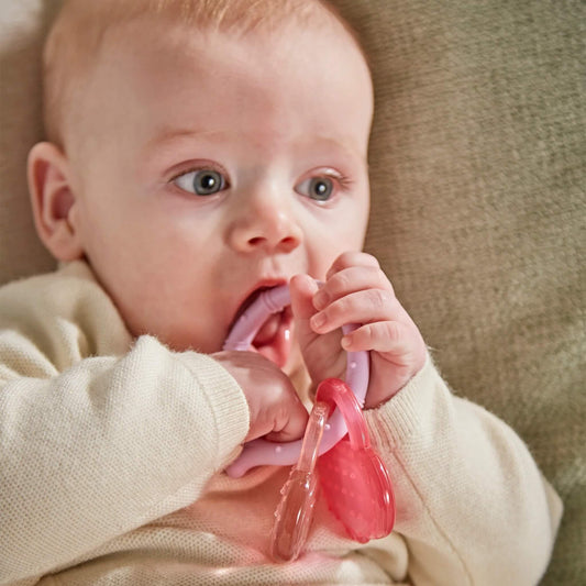 A baby sitting against a soft cushion chewing a lilac silicone teething ring with attached pink and clear monkey face cooling teethers.