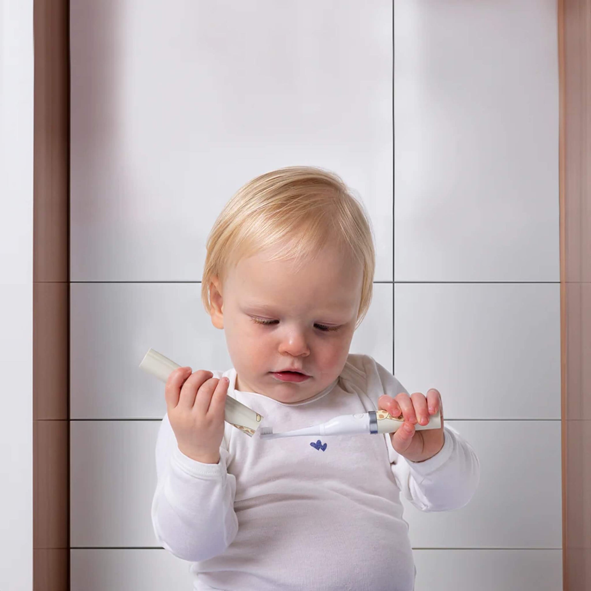 A toddler sitting indoors holding an electric toothbrush handle in one hand and the printed storage tube in the other while examining the parts.