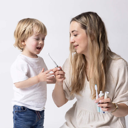 An adult and a young child looking closely at a baby electric toothbrush, with the adult holding toothpaste in the other hand.
