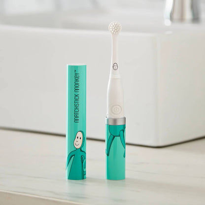 A turquoise monkey-themed baby electric toothbrush standing on a bathroom countertop beside its matching storage tube.