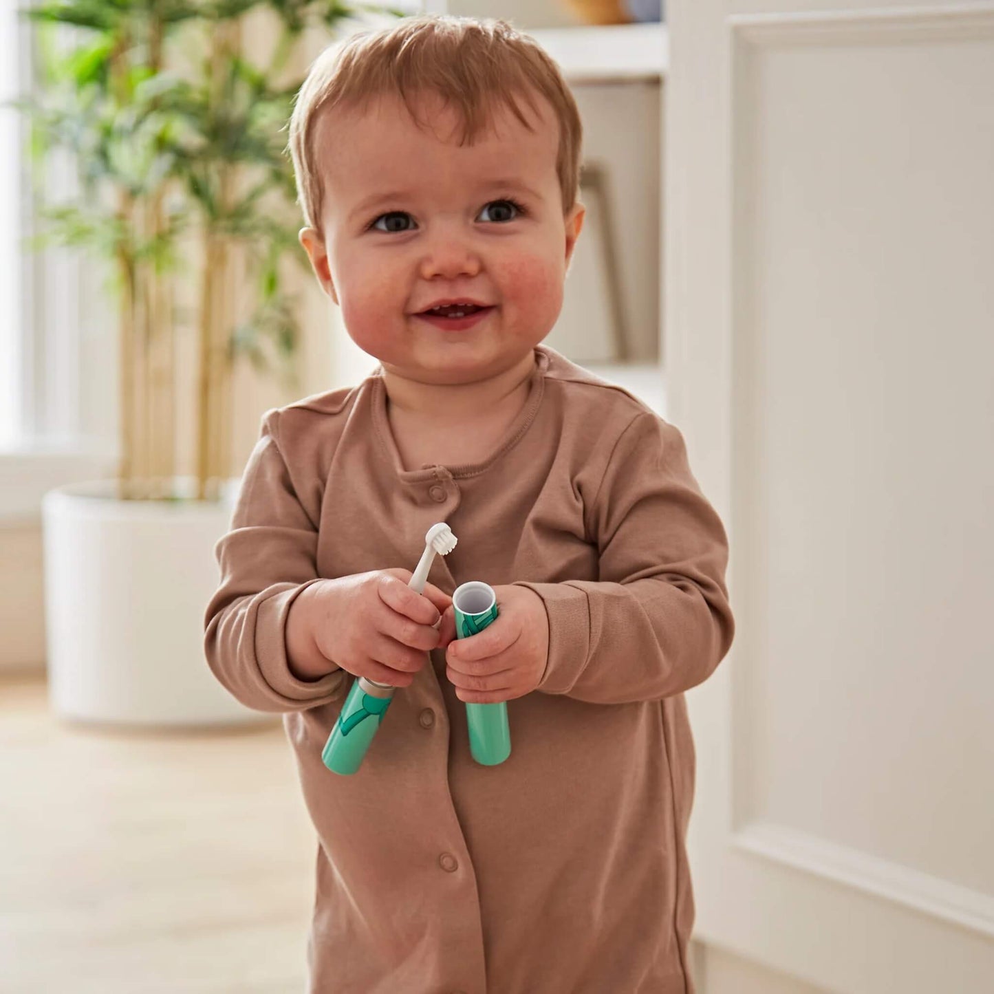 A smiling baby holding a small electric toothbrush and its turquoise storage tube while standing indoors.