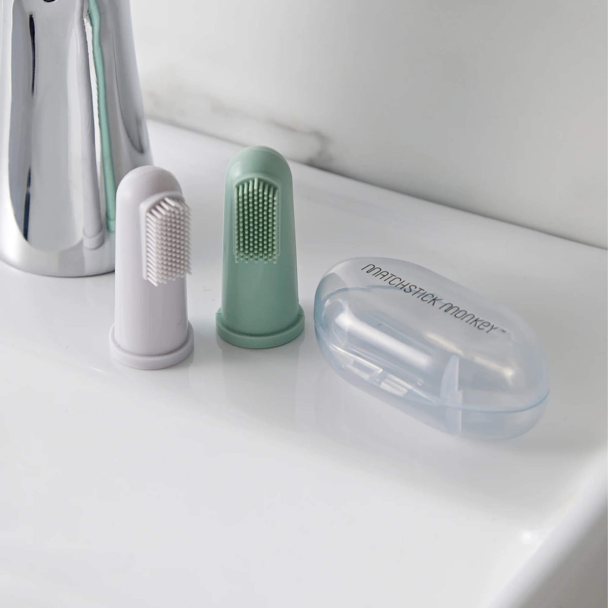 Two silicone baby finger toothbrushes in mint green and grey beside their clear storage case on a bathroom sink.