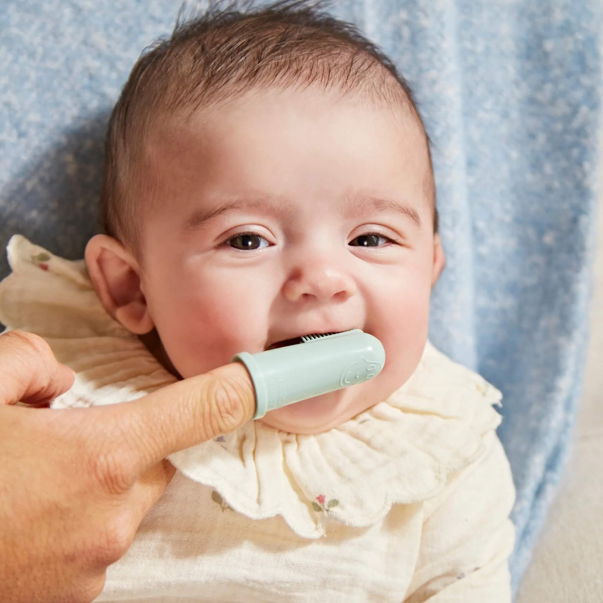 A baby having their first teeth gently brushed with a mint green silicone finger toothbrush.