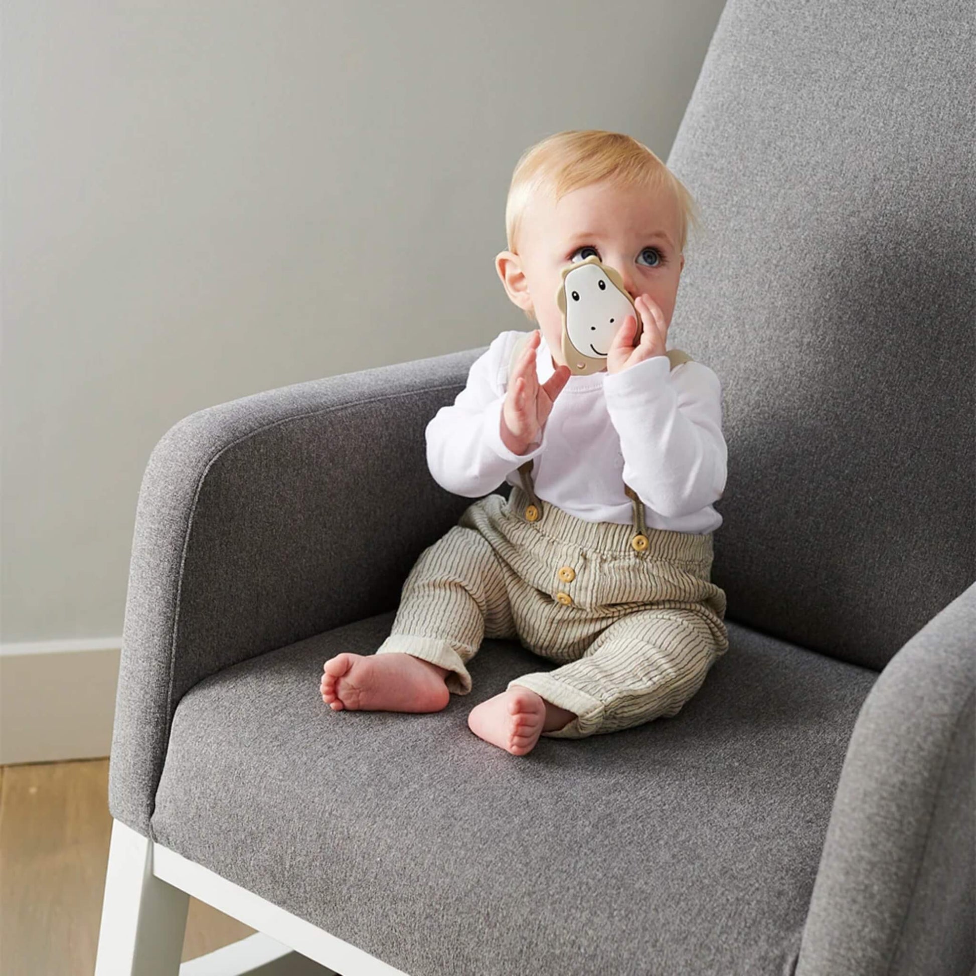 Baby sitting on a grey chair while exploring a flat giraffe-shaped teether with both hands.