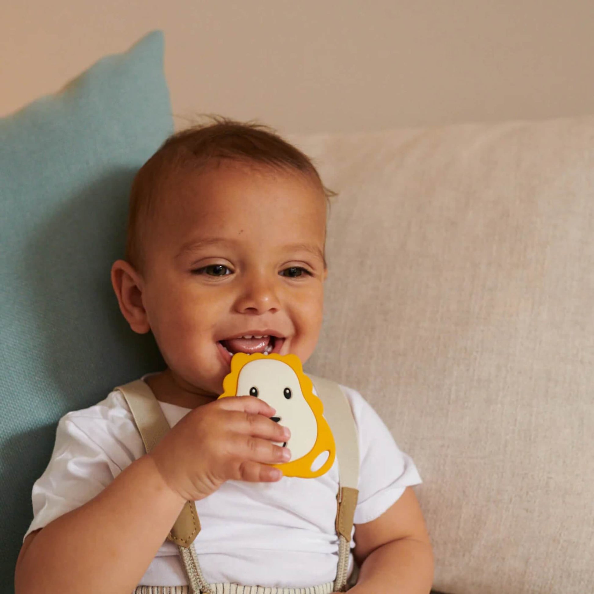 A baby sitting on a sofa holding a lion-shaped flat silicone teether and bringing it towards their mouth.