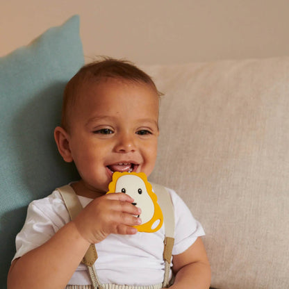 A baby sitting on a sofa holding a lion-shaped flat silicone teether and bringing it towards their mouth.