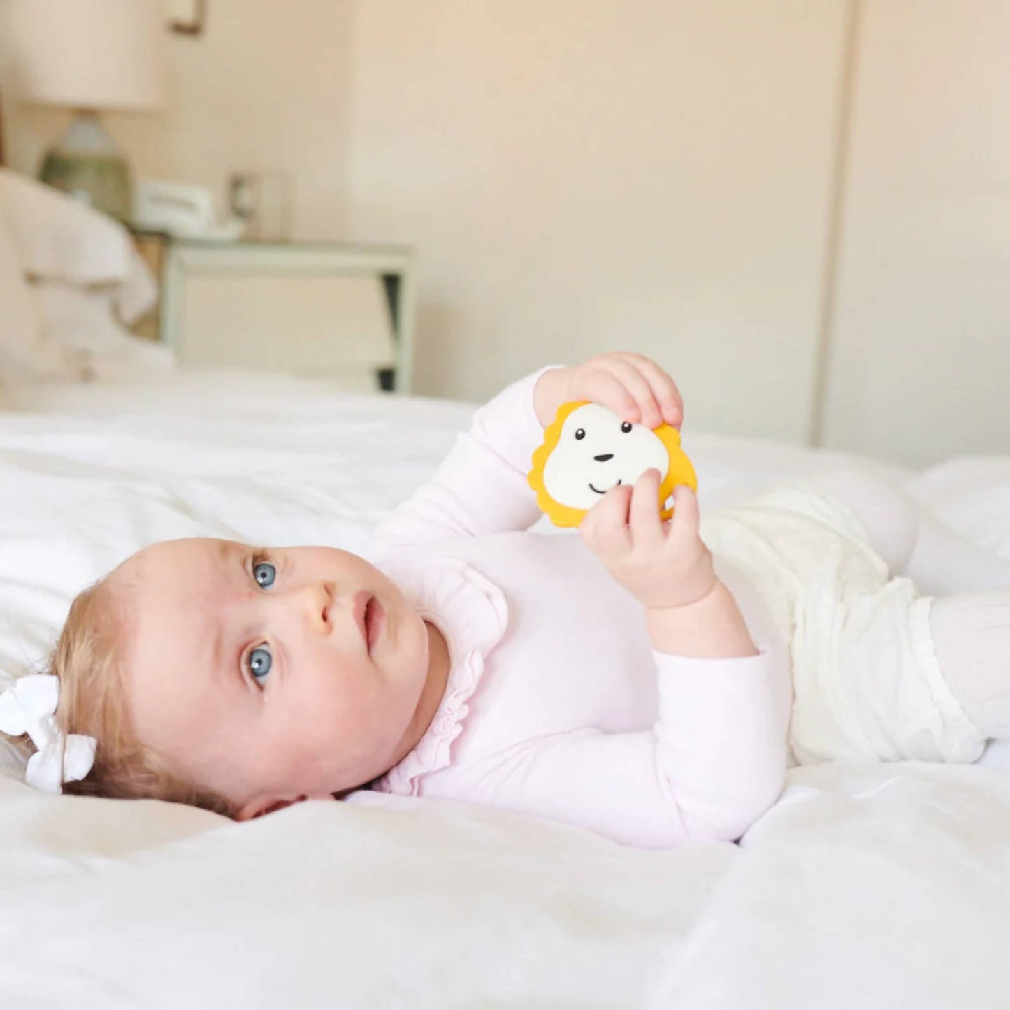 A baby lying on a bed holding a lion-shaped flat silicone teether with both hands while looking upwards.