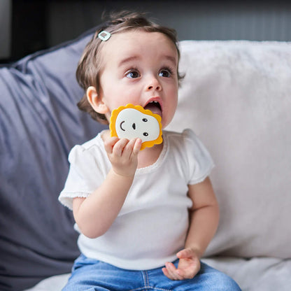 A toddler sitting on a sofa lifting a lion-shaped flat silicone teether towards their mouth.