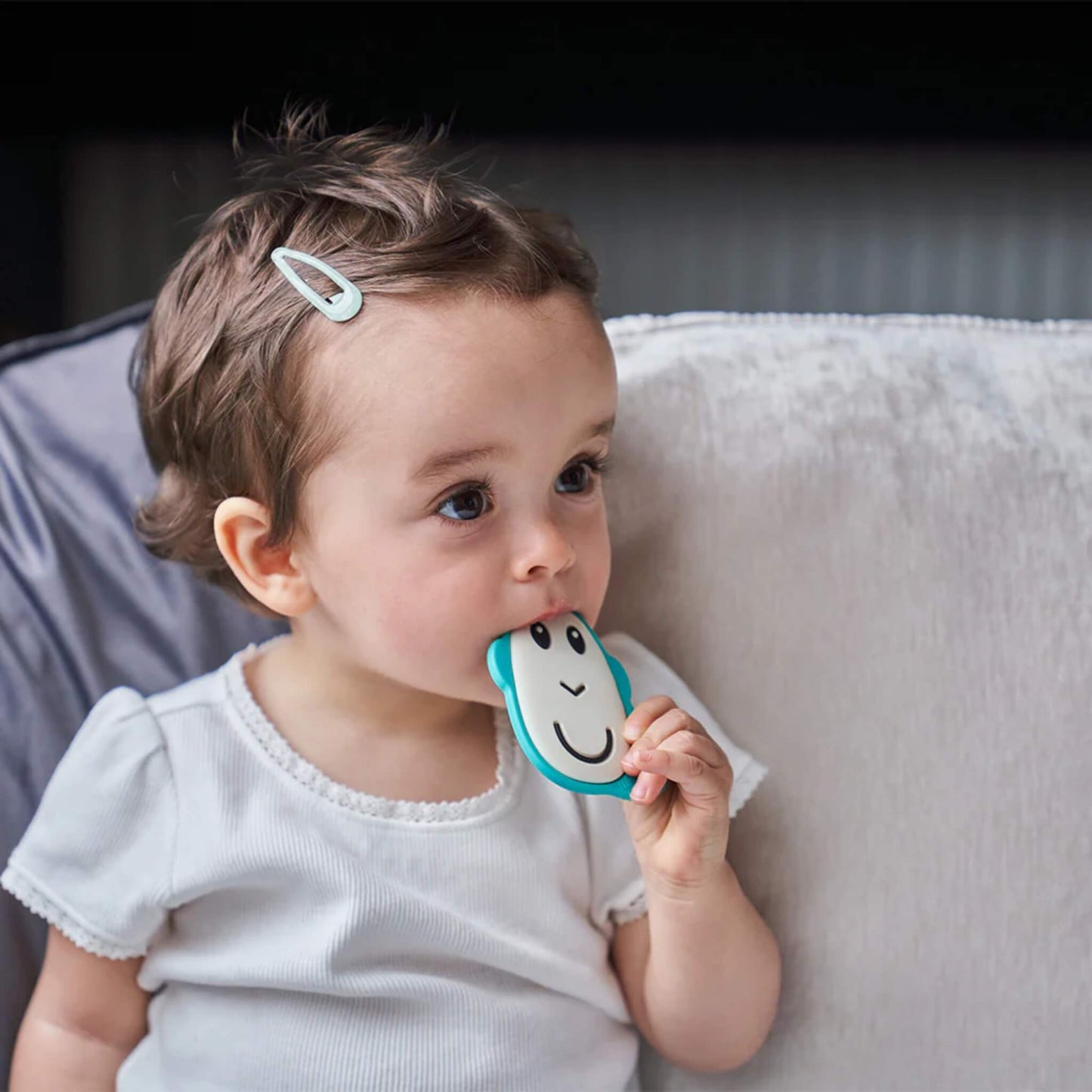 A baby sitting on a sofa, holding a flat monkey-shaped silicone teether and chewing on the smooth front surface.