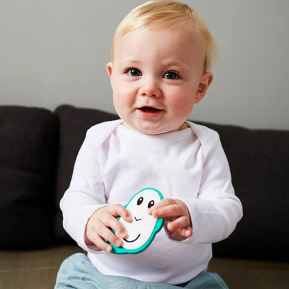 A baby sitting upright on a bed, gripping a flat monkey teether with both hands while looking towards the camera.