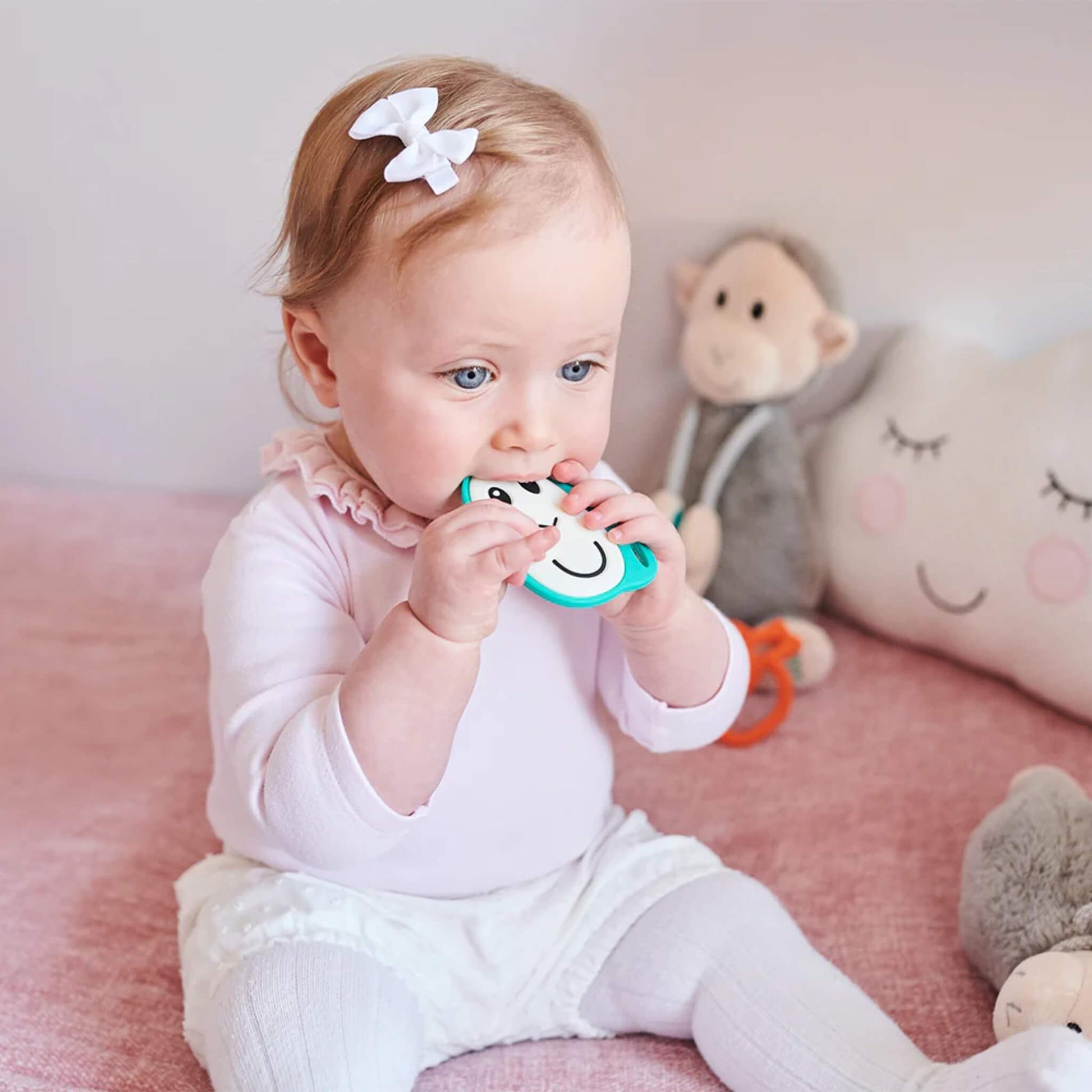 A baby chewing on a flat monkey-shaped silicone teether while sitting in a soft pink nursery space.