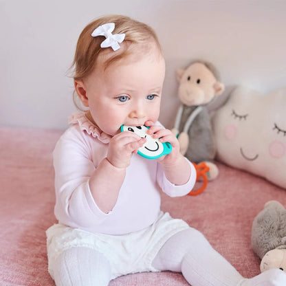 A baby chewing on a flat monkey-shaped silicone teether while sitting in a soft pink nursery space.