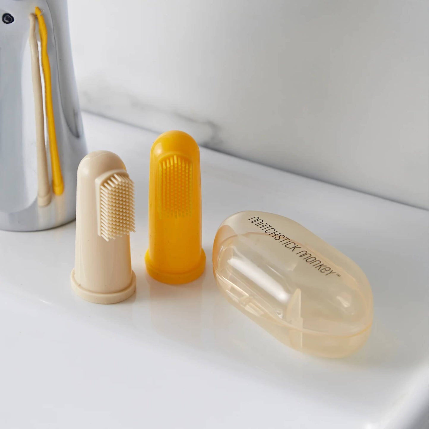 Two silicone finger toothbrushes and protective carry case placed on a bathroom surface beside a toothbrush holder.