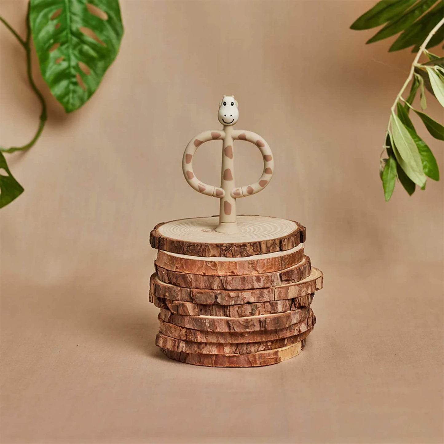 A giraffe-shaped silicone training toothbrush standing on a stack of cut wooden discs against a warm, natural background with green leaves in the frame.