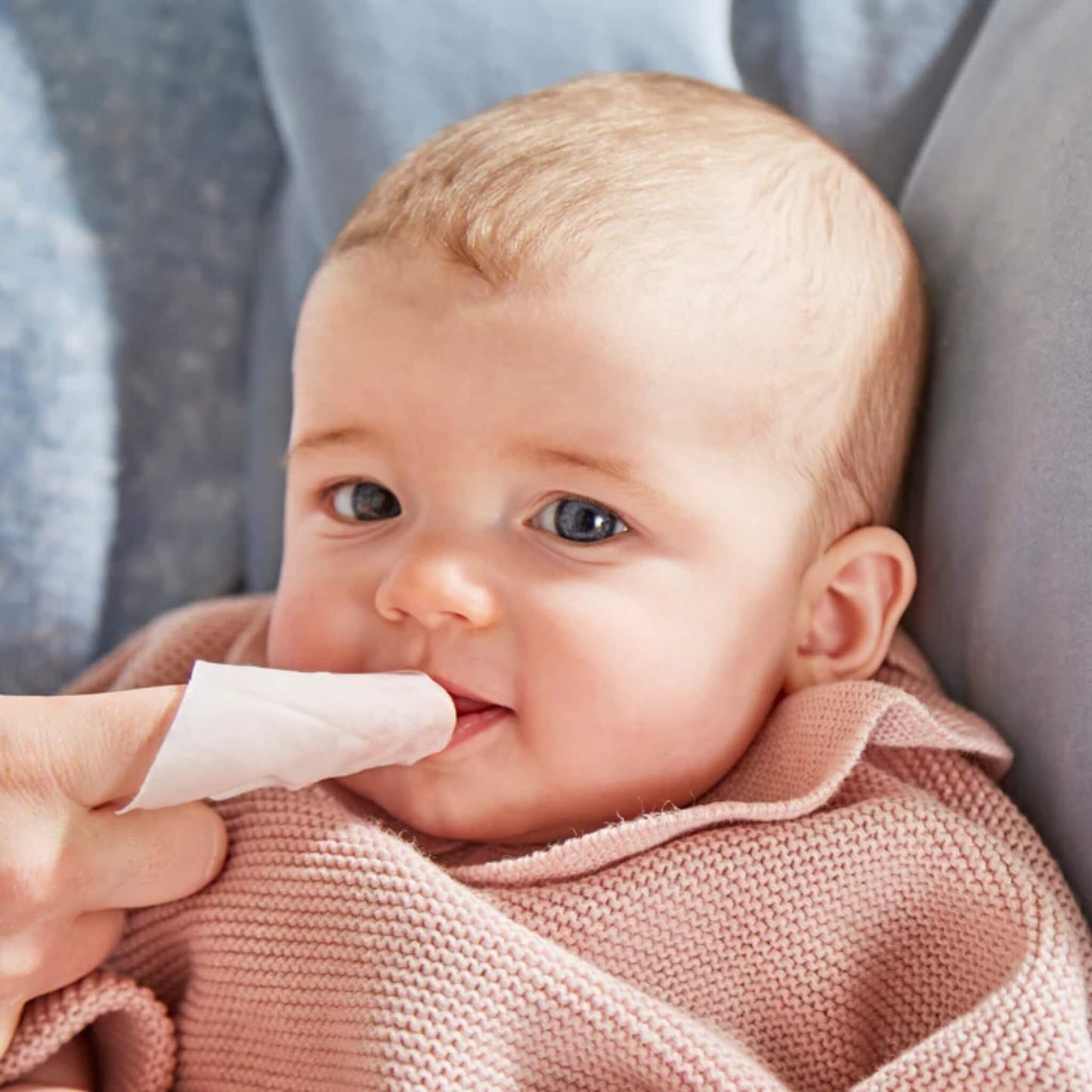 Baby having gums gently cleaned with a soft oral wipe wrapped around an adult finger while sitting in a caregiver’s arms.