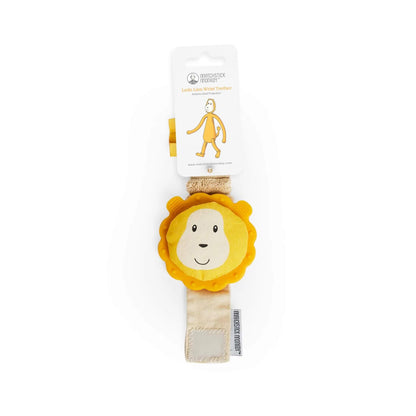 A wrist teether is displayed inside its packaging, with the yellow lion topper visible above the folded fabric strap.