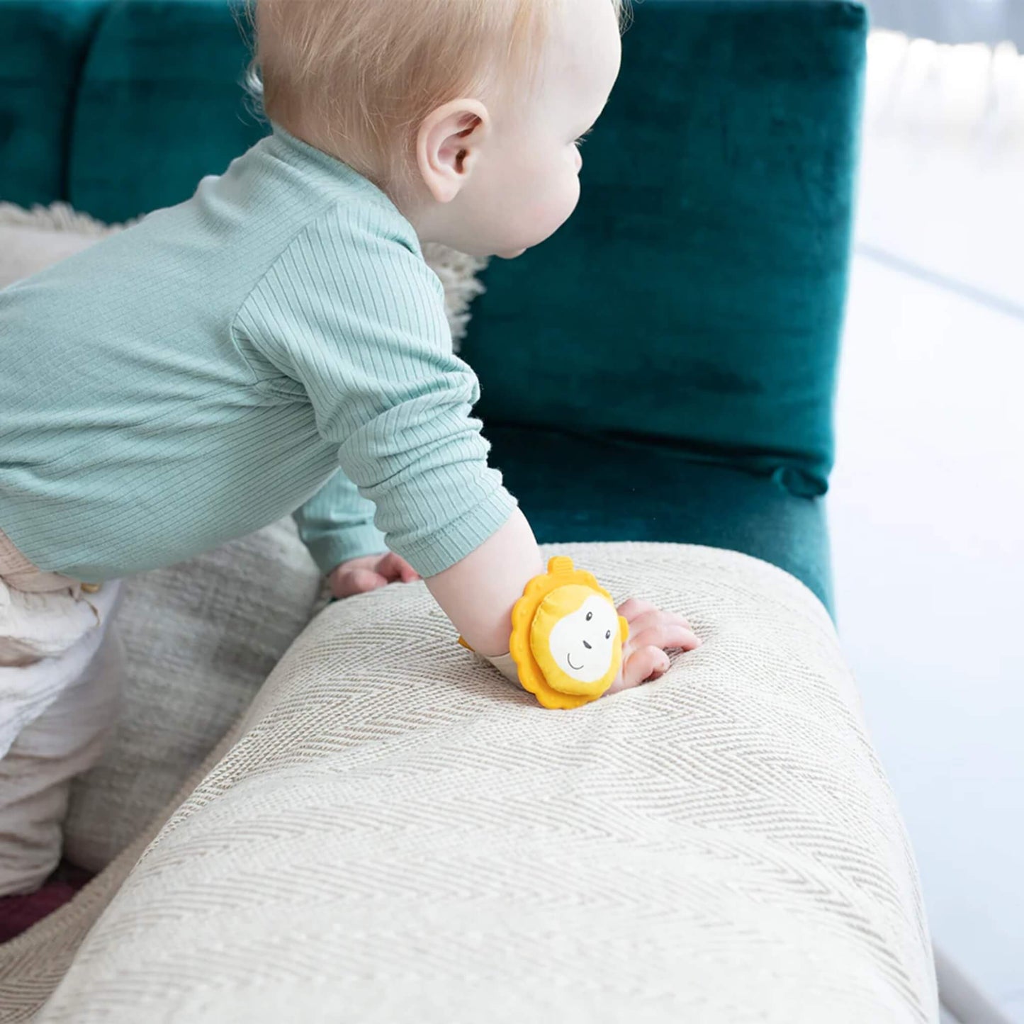 A baby leans forward on a sofa with the wrist teether fastened around their wrist, the yellow lion topper resting against the fabric surface.