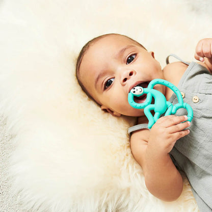 A baby is lying on a fluffy cream rug and chewing on a green monkey-shaped teether, holding it to its mouth.