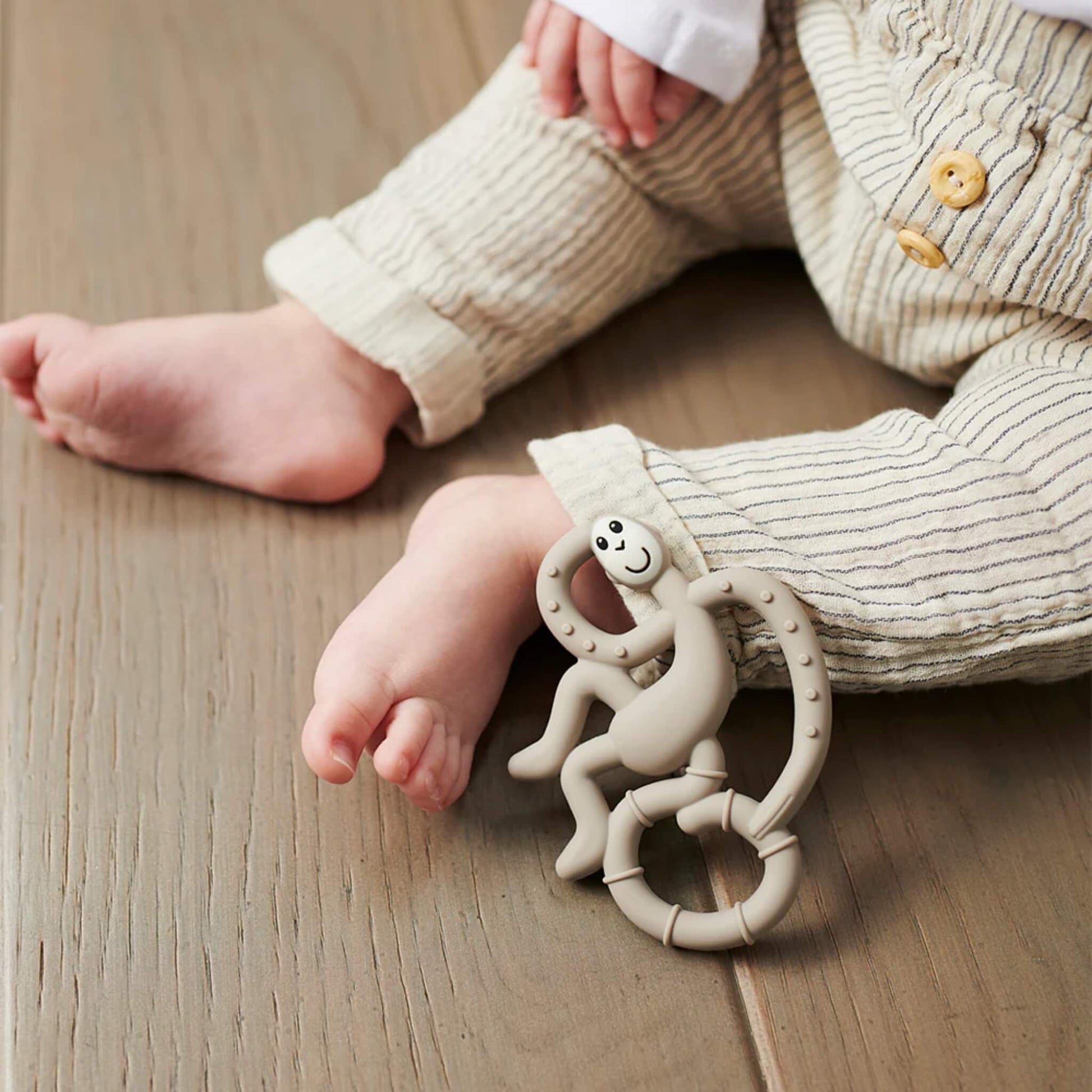 A baby’s lower legs and feet resting on a wooden floor with a grey monkey-shaped teether placed next to the child's ankle.