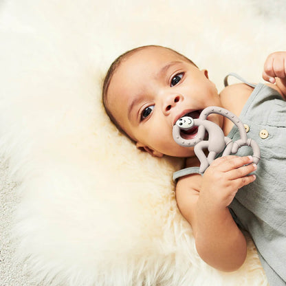 A baby lying on a fluffy cream rug while chewing on a grey monkey-shaped teether held between both hands.