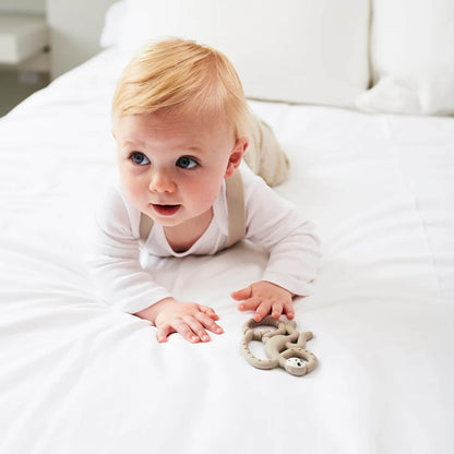 A crawling baby on a white bed reaching forward with one hand while a grey monkey-shaped teether rests near their other hand.