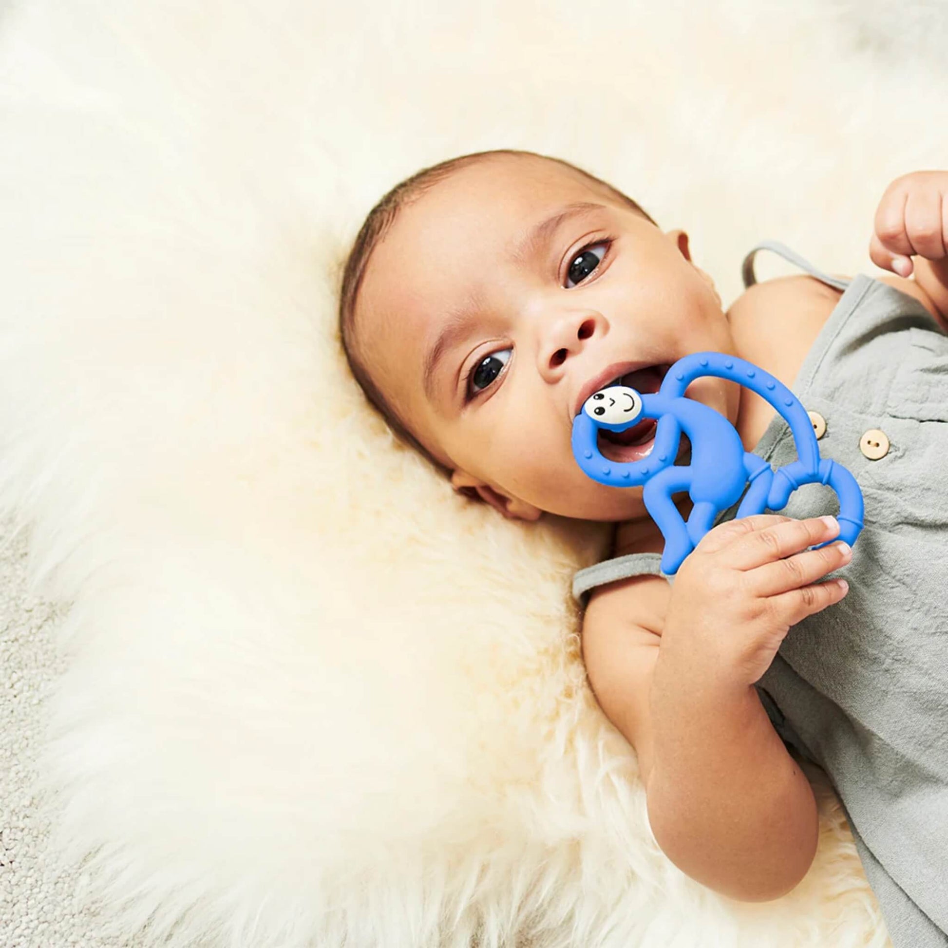 Baby lying on a cream fluffy rug while holding a blue monkey-shaped teether and chewing on one of the textured loops.