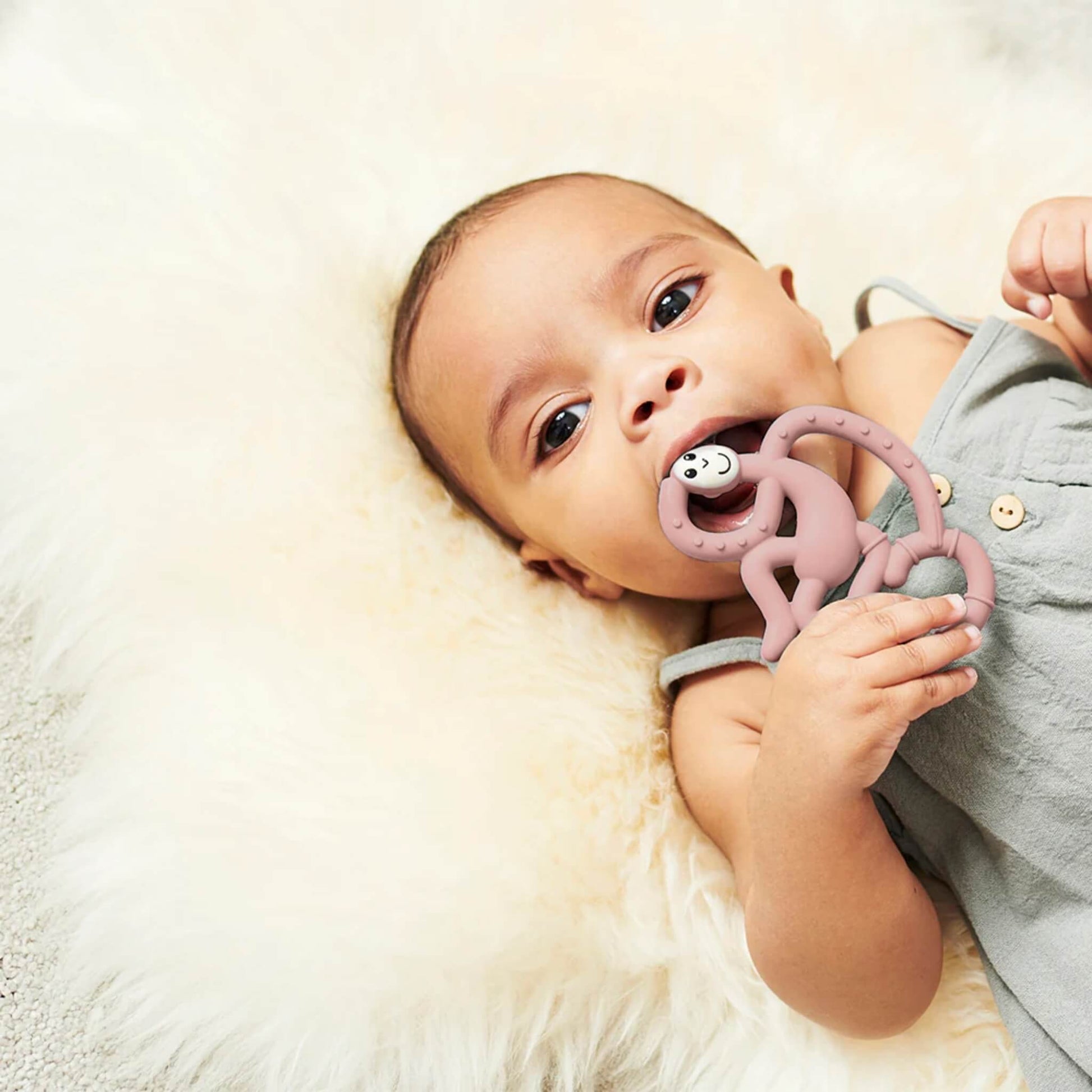 Baby lying on a soft cream blanket holding and chewing a dusty pink monkey-shaped silicone teether.