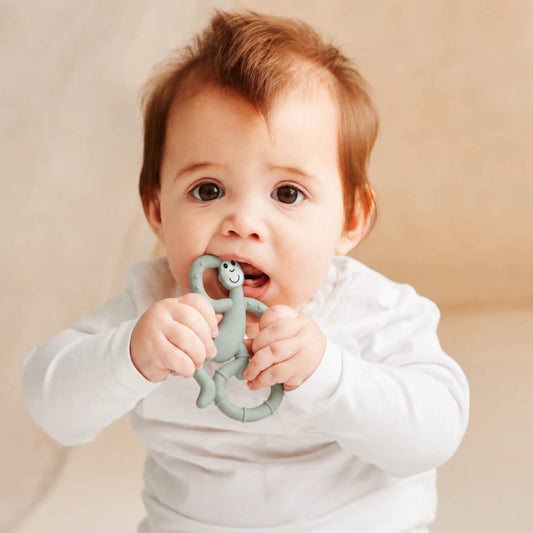 Baby holding and chewing a mint green monkey-shaped silicone teether while wearing a white long-sleeve top.