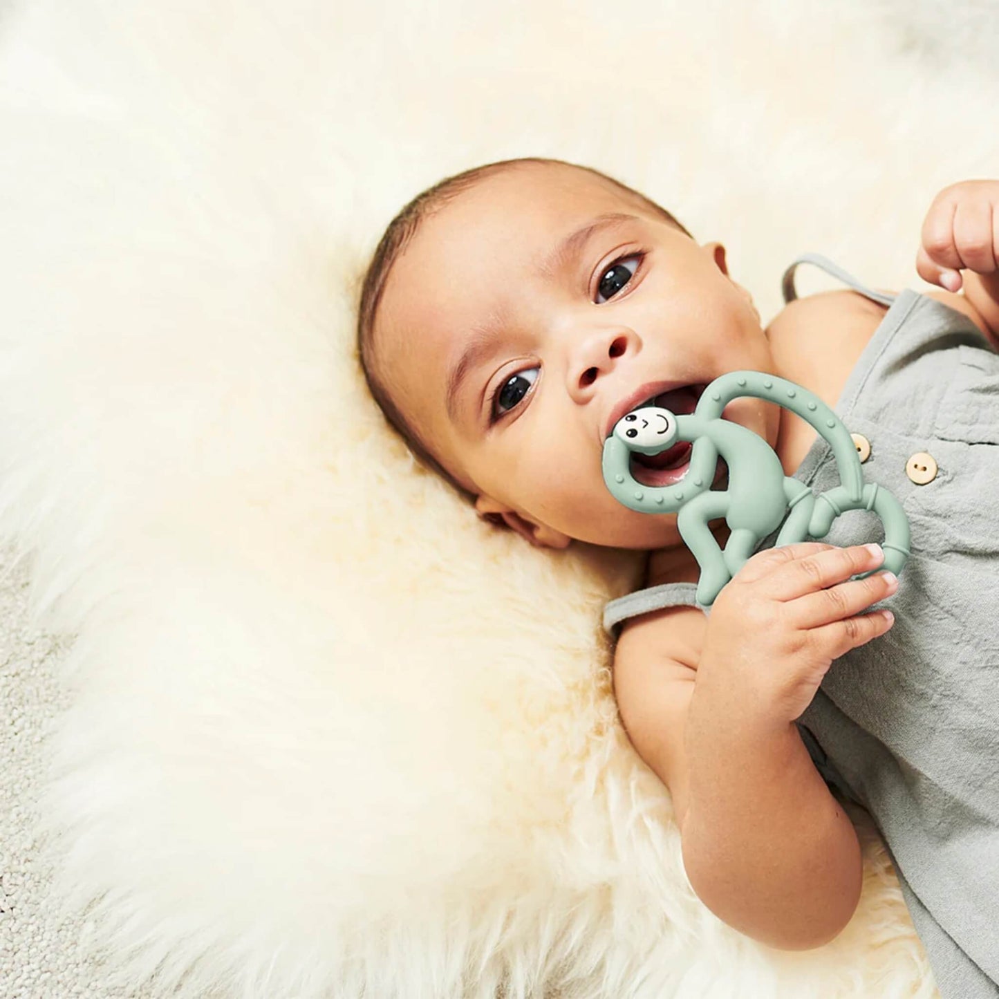 Baby lying on a soft cream blanket holding and chewing a mint green monkey-shaped silicone teether.