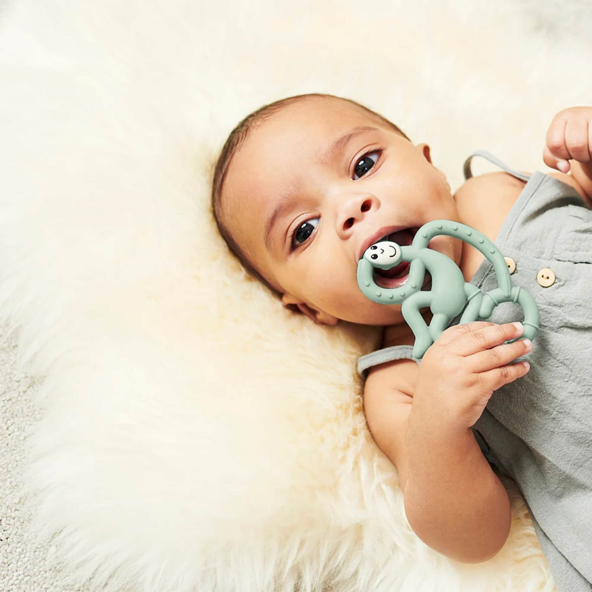 Baby lying on a soft cream blanket holding and chewing a mint green monkey-shaped silicone teether.
