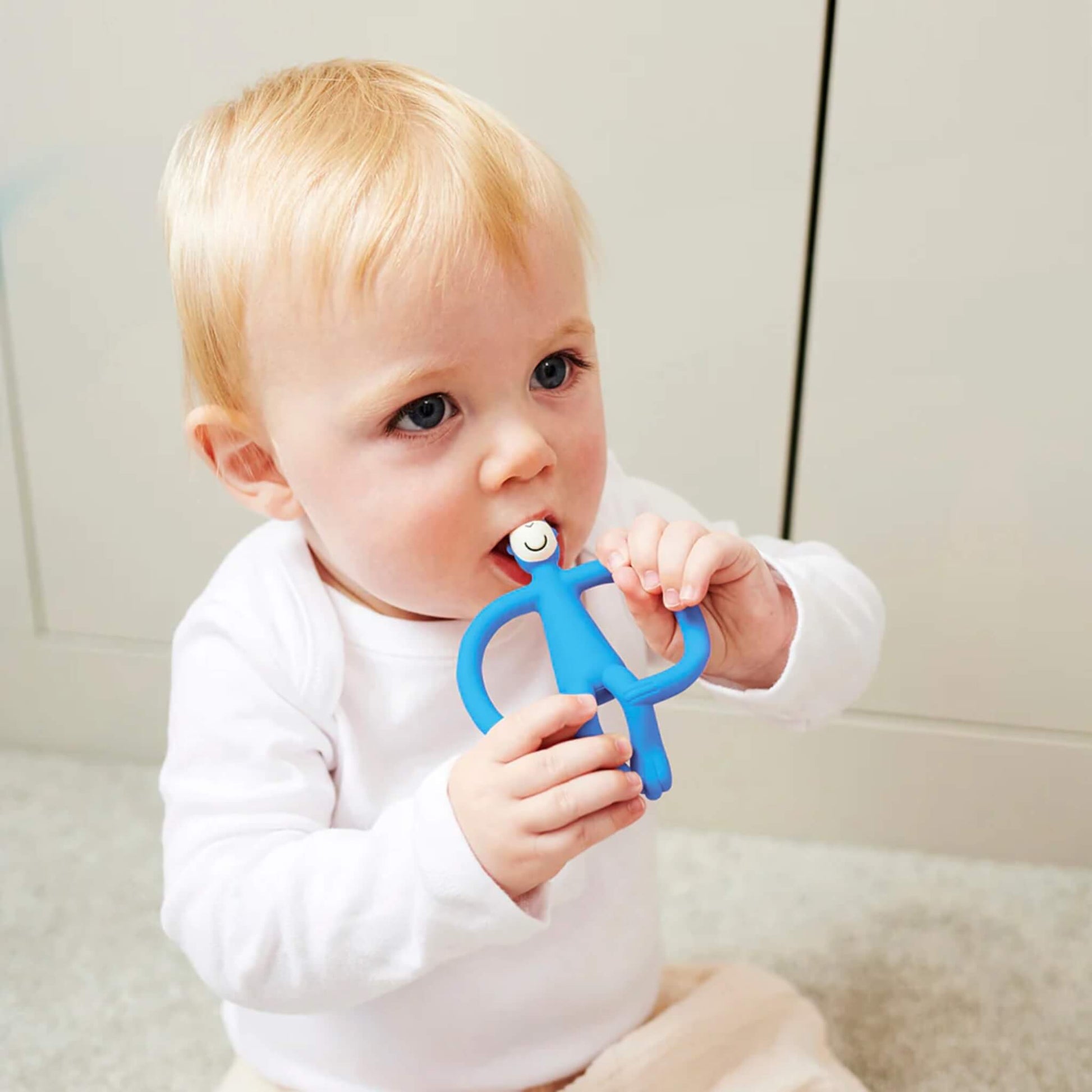 A baby sitting on a soft cream rug holding a blue teether and chewing on the textured head while looking towards the camera.