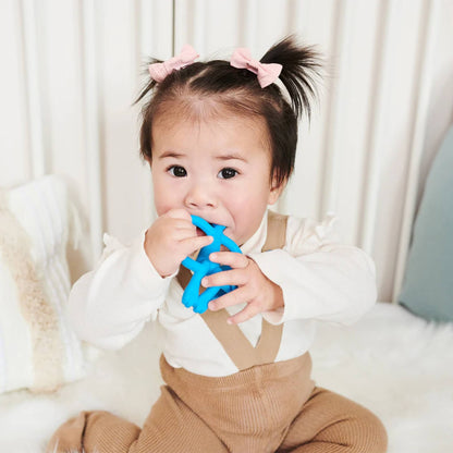 A toddler sitting on a cushioned surface holding a blue teether with both hands and chewing on the top section.