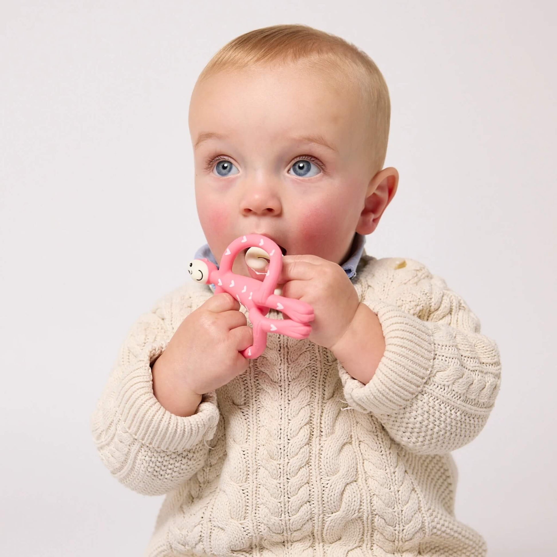 A baby in a cream cable-knit cardigan chewing a pink Matchstick Monkey Original Teether decorated with white hearts.