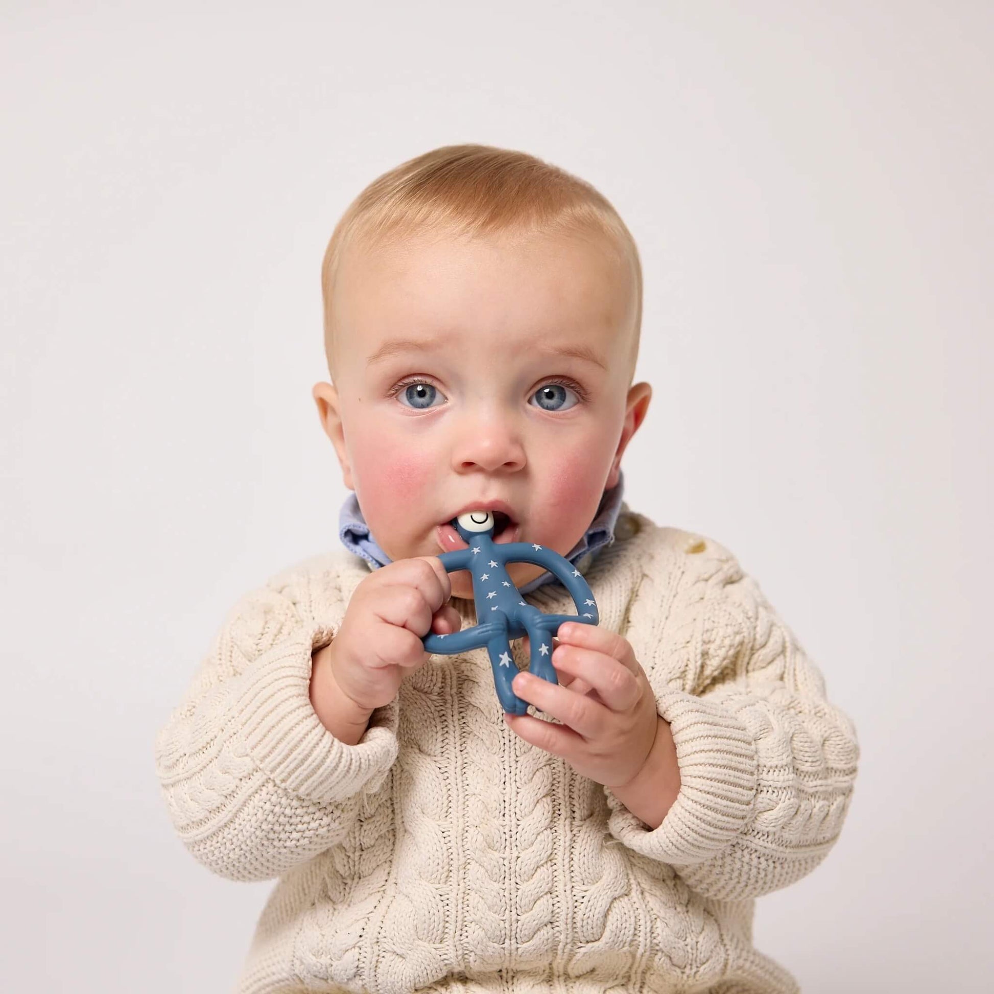 Matchstick Monkey Original Teether with stars design held by a baby wearing a cream knitted cardigan, chewing the easy-grip silicone teether.