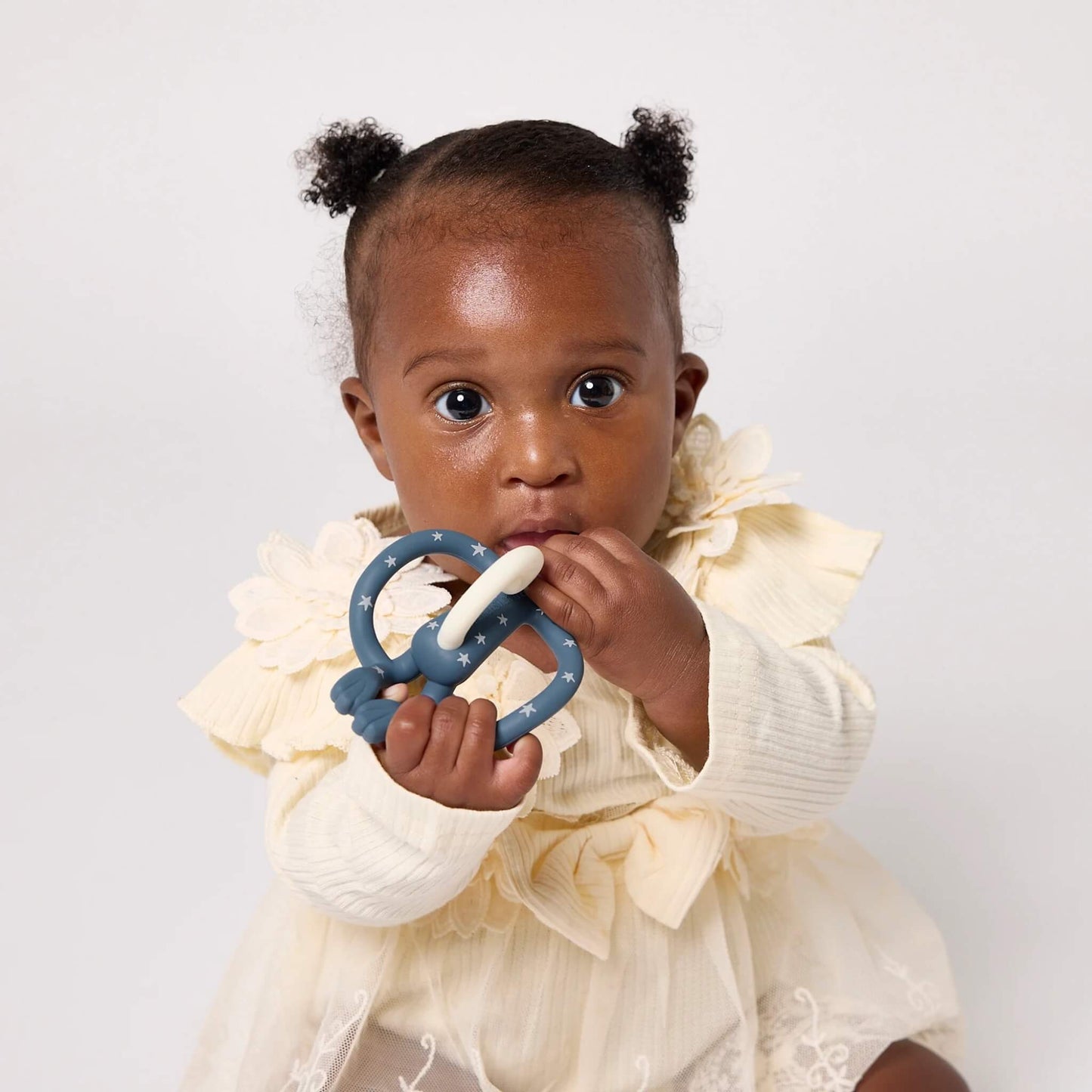 A baby wearing a cream dress holding and chewing a blue star-patterned silicone teether.
