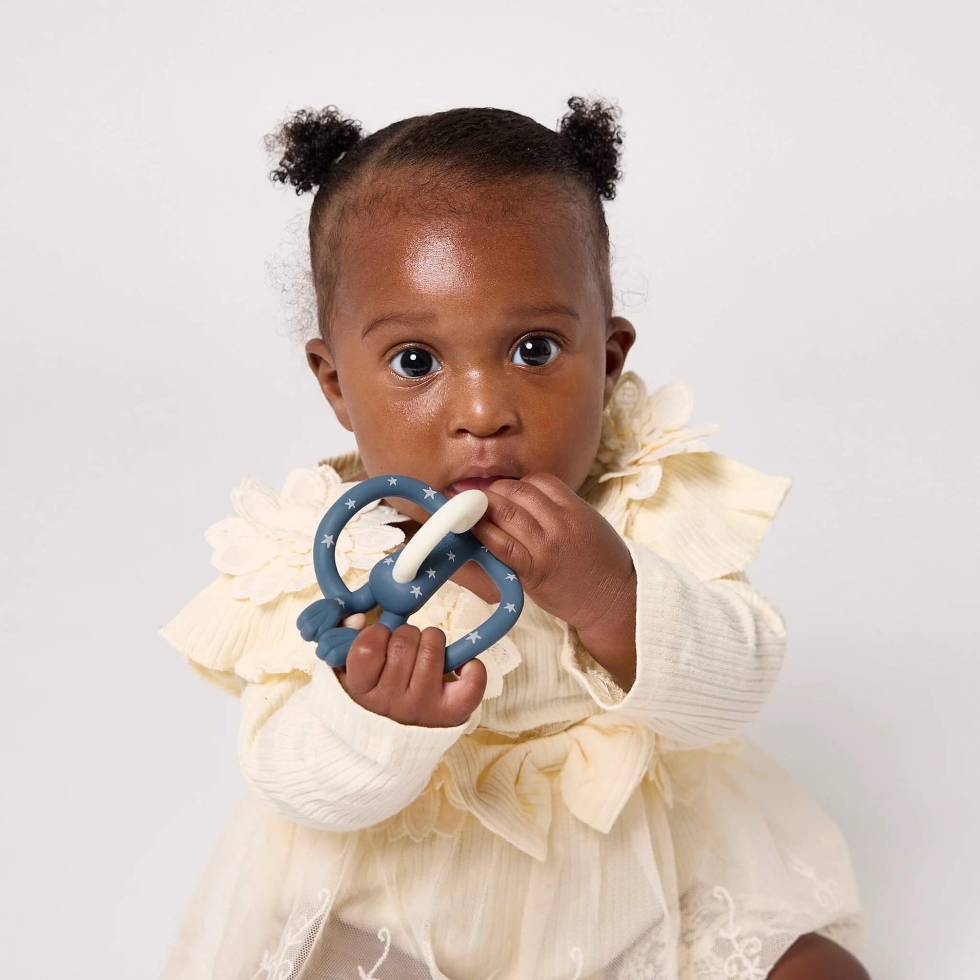 A baby wearing a cream dress holding and chewing a blue star-patterned silicone teether.