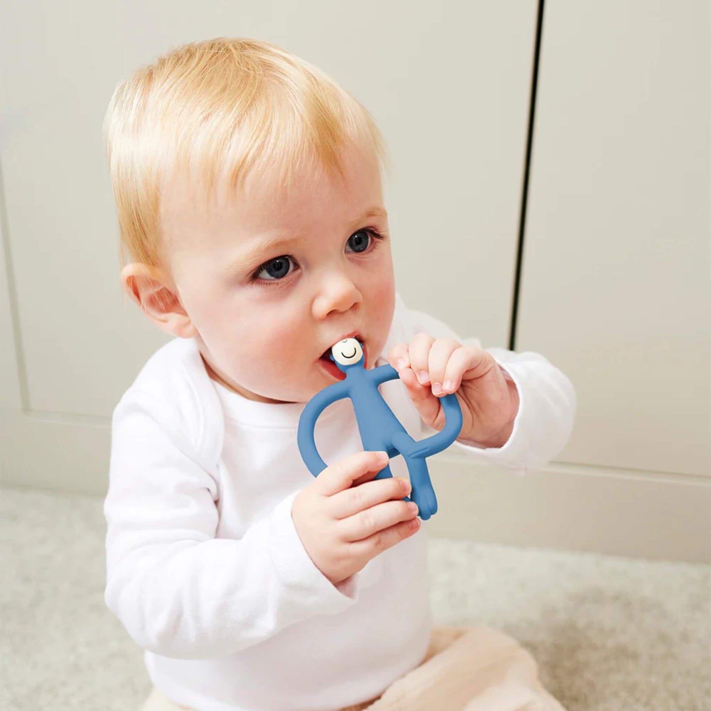A baby sitting on the floor holding a blue silicone monkey teething Toy and biting the textured head while gripping the wide looped arms.